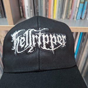 May include: Black baseball cap with the word "Hellripper" embroidered in white gothic lettering. The cap has a curved peak. The background shows a shelf of vinyl records.