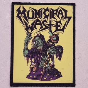 May include: A rectangular patch with a black border and a yellow background. The patch features the text "MUNICIPAL WASTE" in a jagged, stylized font. Below the text is a green and purple illustration of a zombie-like figure holding a staff.