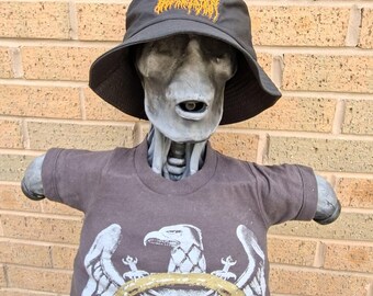 May include: A grey statue wearing a black bucket hat with yellow embroidery and a brown t-shirt. The t-shirt features a white eagle graphic and the word "SLAYER" in orange and black.