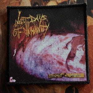 Puede incluir: Parche cuadrado negro con texto dorado "Last Days of Humanity". El diseño abstracto presenta tonos de morado, rojo y blanco. El texto "Putrefaction in progress" también es visible en el parche.