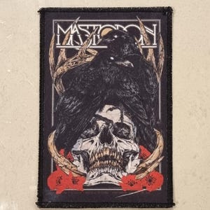 May include: Black rectangular patch featuring the band name "Mastodon" above a design of two black birds, a skull, antlers, and red flowers. The patch has a black border.
