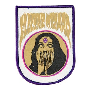 Pode incluir: Patch bordado com borda roxa e fundo branco. O patch apresenta o texto "ELECTRIC WIZARD" em uma fonte dourada estilizada. Abaixo, uma imagem circular de uma mulher com cabelo preto, cobrindo o rosto.