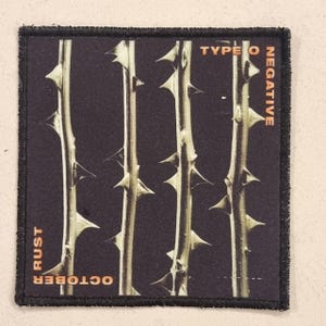 Pode incluir: Um patch quadrado com fundo preto com quatro caules verticais com espinhos. Os caules são de cor castanho claro. As palavras "TYPE O NEGATIVE" e "OCTOBER RUST" são impressas em laranja.
