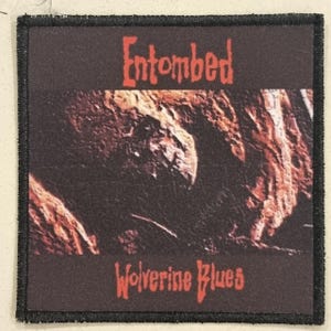 Puede incluir: Un parche cuadrado con borde negro y fondo oscuro. El parche presenta el nombre de la banda "Entombed" en rojo, sobre el texto "Wolverine Blues" en rojo. El centro muestra una imagen borrosa y texturizada.