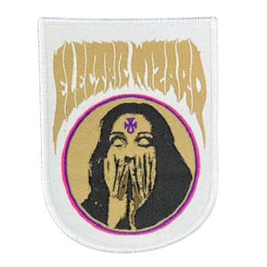 May include: A white patch with a gold and purple design. The patch features the words "ELECTRIC WIZARD" above a circular image of a person with hands covering their face. The person has long black hair and a purple symbol on their forehead.
