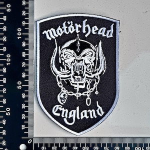 May include: A shield-shaped embroidered patch with the Motörhead logo, a skull with tusks and chains, in white on a black background. The text "Motörhead" and "England" are also in white.