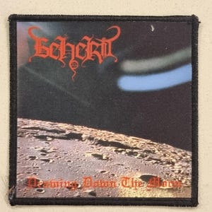 May include: Square patch featuring the band name "Beherit" in red gothic lettering above a lunar landscape. The patch includes the text "Washing Down The Moon" in red, set against a black background with a blue and white glow.