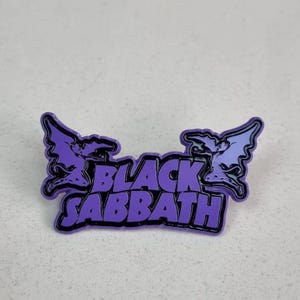 May include: A purple and black enamel pin featuring the words "BLACK SABBATH" in a stylized font. The pin includes two stylized bat-like figures on either side of the text, with a white background.