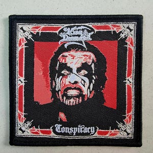 Può includere: Toppa ricamata nera e rossa con un'immagine bianca di un viso maschile con la barba e il testo "King Diamond" e "Conspiracy".