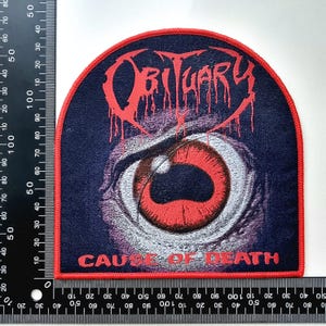 May include: Embroidered patch featuring the word "Obituary" in red dripping letters above a red-eyed illustration. The text "Cause of Death" is below the eye. The patch has a navy blue background and a red border.