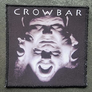 Könnte beinhalten: Quadratischer Aufnäher mit dem Wort "CROWBAR" in weißen Blockbuchstaben oben. Der Aufnäher zeigt ein Graustufenbild eines verzerrten Gesichts mit mehreren Ausdrücken. Der Aufnäher hat einen schwarzen Rand.