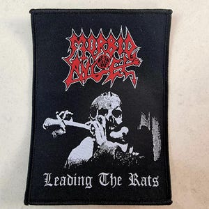 以下が含まれることがあります： 赤文字で「Morbid Angel」というバンド名が書かれた、黒と白の刺繍パッチ。パッチには、ナイフを持った頭蓋骨と「Leading The Rats」という文字が描かれている。