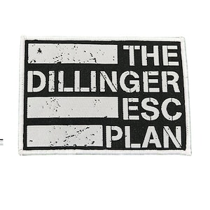 May include: A rectangular black and white patch featuring the text "THE DILLINGER ESC PLAN". The text is in a bold, sans-serif font. The patch has a white border and a distressed appearance.