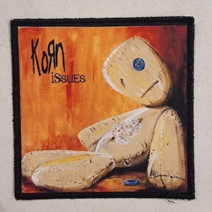 May include: Square patch featuring the word "Korn" above "Issues" in black lettering. The patch depicts a beige, stitched doll against an orange background. A blue button is on the doll's head, and a blue ring is at the bottom.