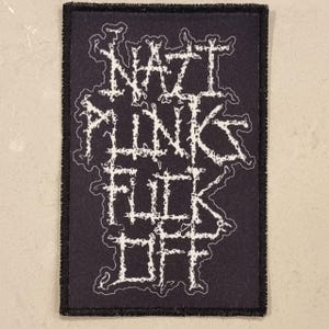 以下が含まれることがあります： 黒い長方形のパッチ。白い文字で「NAZI PUNK FUCK OFF」と書かれており、様式化されたギザギザのフォントです。パッチには黒い縁取りがあり、おそらく布製です。