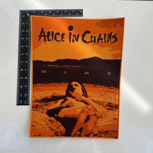Pode incluir: Remendo retangular laranja com o texto "Alice in Chains" e a palavra "Dirt". O remendo apresenta uma ilustração de uma pessoa. O remendo tem aproximadamente 30 cm de altura e 20 cm de largura.