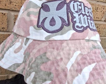 May include: A camouflage bucket hat in pink, white, and grey. The hat has an embroidered patch with a cross design and the text "Clever Miss". The hat is made of a soft, durable fabric.