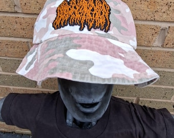 May include: A camouflage bucket hat in shades of pink, white, and grey, with an orange embroidered design. The hat is worn by a mannequin wearing a brown t-shirt.