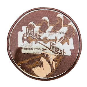 May include: A round, brown patch featuring a stylized razor blade design with the words "Judas Priest" and "British Steel" in white. The patch has a brown border and a tan and brown background.