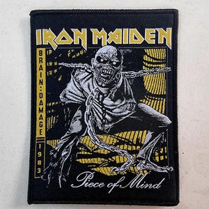 May include: Black and white embroidered patch featuring the band name "Iron Maiden" in gold lettering. The patch depicts a skeletal figure with a chain around its neck and the text "Piece of Mind" in white cursive lettering. The patch also includes the text "Brain Damage 1983" in yellow lettering.
