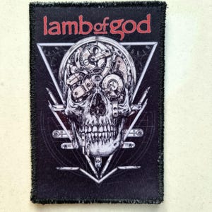 Può includere: Toppa nera con un design dettagliato di teschio con elementi di orologeria e il testo rosso "lamb of god". Il design è incorniciato in una forma geometrica, per i fan della musica metal.
