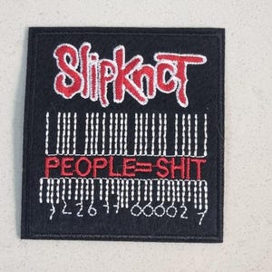 Pode incluir: Patch quadrado preto com o nome da banda "Slipknot" em vermelho, com letras estilizadas. Abaixo, um design de código de barras e a frase "PEOPLE=SHIT" em vermelho. Detalhes e números bordados em branco.
