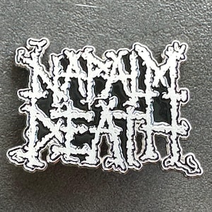 Puede incluir: Un pin de metal negro y blanco con las palabras "NAPALM DEATH" en una fuente estilizada.