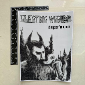 Puede incluir: Un parche rectangular en blanco y negro con el texto "ELECTRIC WIZARD" y "dopethrone". La imagen muestra una figura con cuernos y barba, con un estilo gótico oscuro. Una regla se coloca junto al parche, midiendo en centímetros.