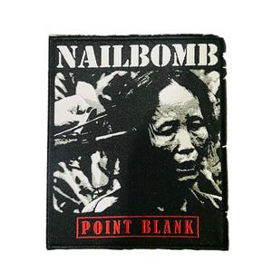 Pode incluir: Um patch retangular em preto e branco com o texto "NAILBOMB" na parte superior e "POINT BLANK" em vermelho na parte inferior. O patch apresenta uma imagem em tons de cinza do rosto de uma pessoa.