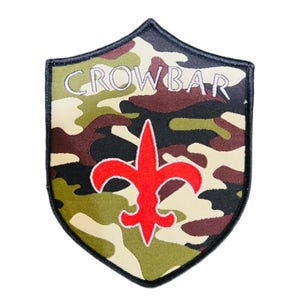 May include: A shield-shaped patch with a camouflage pattern in green, brown, and beige. The word "CROWBAR" is embroidered in white across the top. A red fleur-de-lis design is centered below the text, outlined in white.