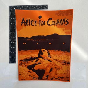 Pode incluir: Patch retangular laranja e castanho com o nome da banda "Alice in Chains" e a palavra "Dirt". A imagem mostra uma pessoa em uma paisagem desértica. O patch é retangular.