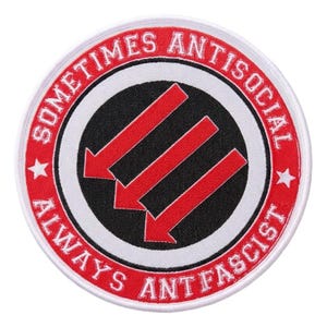 Puede incluir: Parche redondo con borde blanco, texto rojo y centro negro. El texto dice "SOMETIMES ANTISOCIAL ALWAYS ANTIFASCIST". Tres flechas rojas apuntan hacia abajo en el centro. Dos estrellas blancas están en el borde rojo.