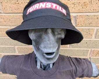 May include: Black bucket hat with the word "TURNSTILE" in red and white lettering. The hat is worn by a grey statue wearing a brown t-shirt.