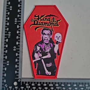 Könnte beinhalten: Roter Sarg-förmiger Aufnäher mit dem Text "King Diamond" in Gelb und Schwarz und einem Fledermaus-Motiv. Der Aufnäher zeigt eine Figur, die einen Schädel und ein Kreuz hält. Der Aufnäher ist etwa 10 cm hoch.