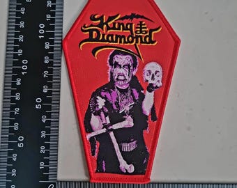 May include: Red coffin-shaped patch featuring the text "King Diamond" in yellow and black, with a bat graphic. The patch depicts a figure holding a skull and a cross. The patch is approximately 4 inches tall.