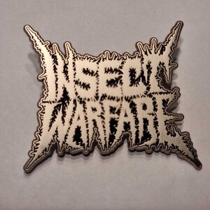 Puede incluir: Un pin de metal plateado con un diseño en blanco y negro. El pin presenta las palabras "Insect Warfare" en una fuente estilizada.