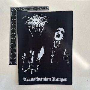 May include: Black rectangular patch featuring a white graphic of a screaming figure and a band logo. The text "Transilvanian Hunger" is printed in white at the bottom. A ruler is placed next to the patch for scale.
