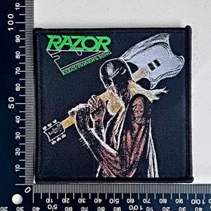 Może przedstawiać: Czarno-zielona naszywka haftowana z nazwą zespołu "RAZOR" w zielonych literach i tekstem "EXECUTIONER'S SONG" poniżej. Naszywka przedstawia czarno-białą ilustrację osoby trzymającej gitarę.