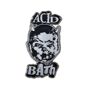 May include: A black and white enamel pin featuring a devil-faced baby design. The pin has the words "ACID" above the baby's head and "BATH" below. The pin is set against a white background.