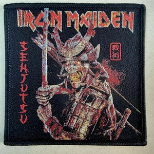 May include: Black embroidered patch with the band name "IRON MAIDEN" in red letters. The patch features a red and gold samurai warrior with a sword, and the Japanese text "SENJUTSU" in red.
