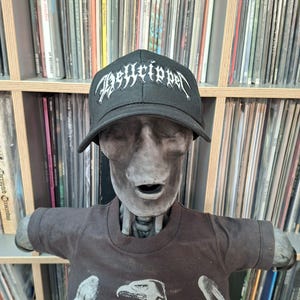 May include: A black baseball cap with the word "Desyripper" in white gothic lettering. The cap is worn by a mannequin wearing a dark brown t-shirt. The background features a shelf filled with vinyl records.