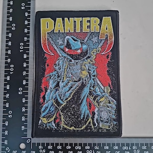 May include: Black rectangular patch featuring the word "PANTERA" in gold and a skeletal figure in a cowboy hat. The figure is holding a bottle and is set against a red and blue background.
