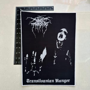 Puede incluir: Parche en blanco y negro con el logotipo de la banda Darkthrone y una ilustración de una persona gritando. El texto "Transilvanian Hunger" se muestra en la parte inferior. El parche es rectangular.