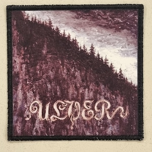 May include: Square patch with a black border featuring a landscape of a mountain with trees and a cloudy sky. The word "ULVER" is written in a decorative font across the bottom of the patch.