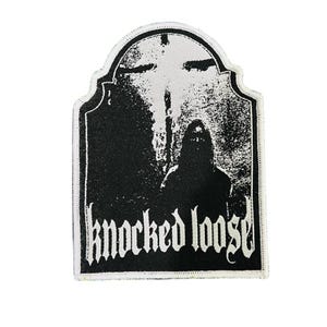 Könnte beinhalten: Schwarz-weißer Aufnäher mit einer Silhouette und einem Kreuz, mit dem Schriftzug "knocked loose" in gotischen Buchstaben. Der Aufnäher hat einen weißen Rand und eine rechteckige Form mit gewölbter Oberseite.