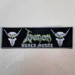 May include: Black rectangular patch embroidered with the word "Venom" in a stylized font, with a green and silver outline. Below the word "Venom" is the text "BLACK METAL". The patch features a skull on each side.