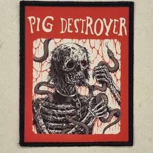 Op de afbeelding: Een rechthoekige patch met een rode rand en zwarte rand. De patch heeft de tekst "PIG DESTROYER" in het wit. De afbeelding toont een skeletfiguur met slangen en rode ogen. De achtergrond is wit met rode druppels.