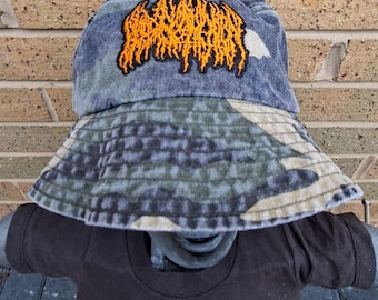 May include: A camouflage bucket hat with a dark orange embroidered design. The hat is made of a durable fabric and has a wide brim. The hat is displayed on a black t-shirt.