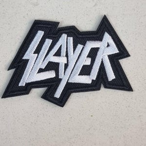 May include: A black and white embroidered patch with the word "SLAYER" in a bold, stylized font. The patch has a black border and a jagged, angular shape. The text is in white.