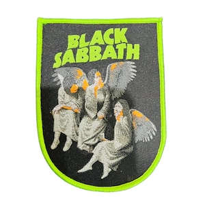 Puede incluir: Parche de Black Sabbath con forma de escudo. El parche presenta el nombre de la banda en letras verdes neón sobre un gráfico de tres figuras aladas. El parche tiene un fondo negro y un borde verde neón.